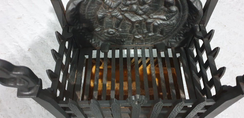 A compact black cast iron fire grate with picket-style front bars, ornamental finials, and a relief-cast backplate depicting figures. View VI
