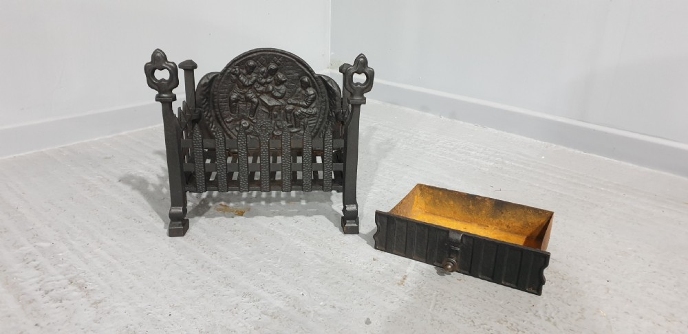 A compact black cast iron fire grate with picket-style front bars, ornamental finials, and a relief-cast backplate depicting figures. View VII