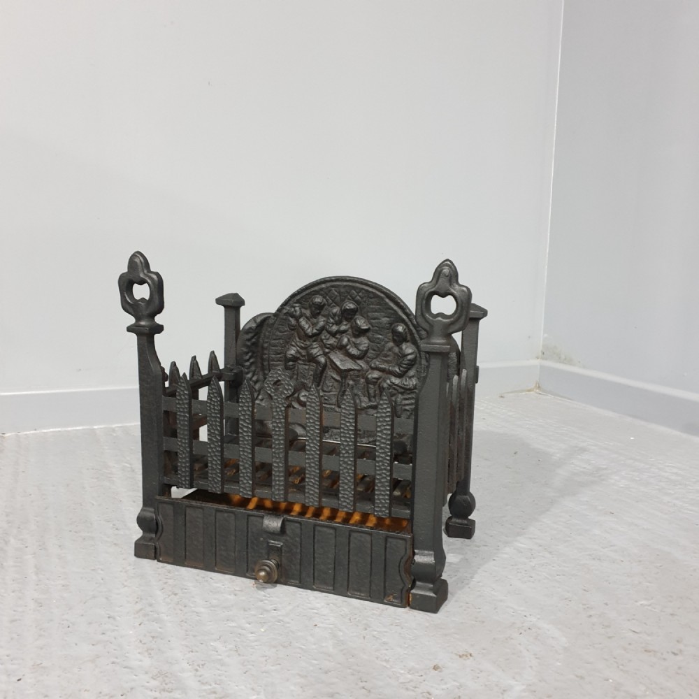 A compact black cast iron fire grate with picket-style front bars, ornamental finials, and a relief-cast backplate depicting figures.