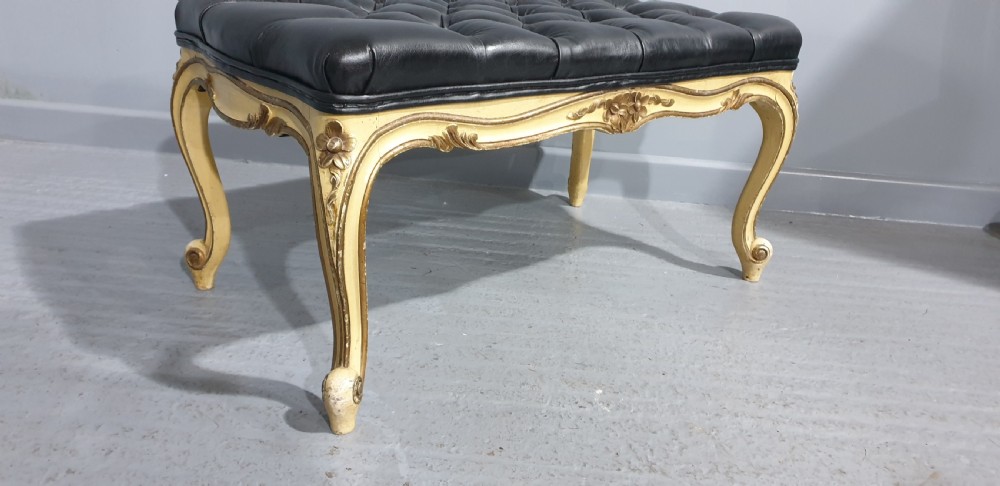 A long rectangular stool featuring a black buttoned leather tufted seat set upon a carved cream and gold painted wooden frame with cabriole legs. View V