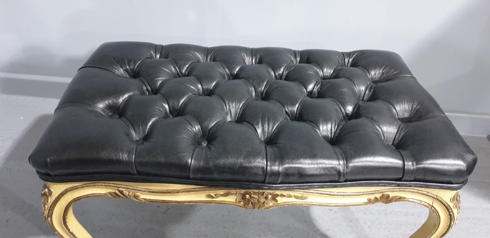 A long rectangular stool featuring a black buttoned leather tufted seat set upon a carved cream and gold painted wooden frame with cabriole legs. View VI