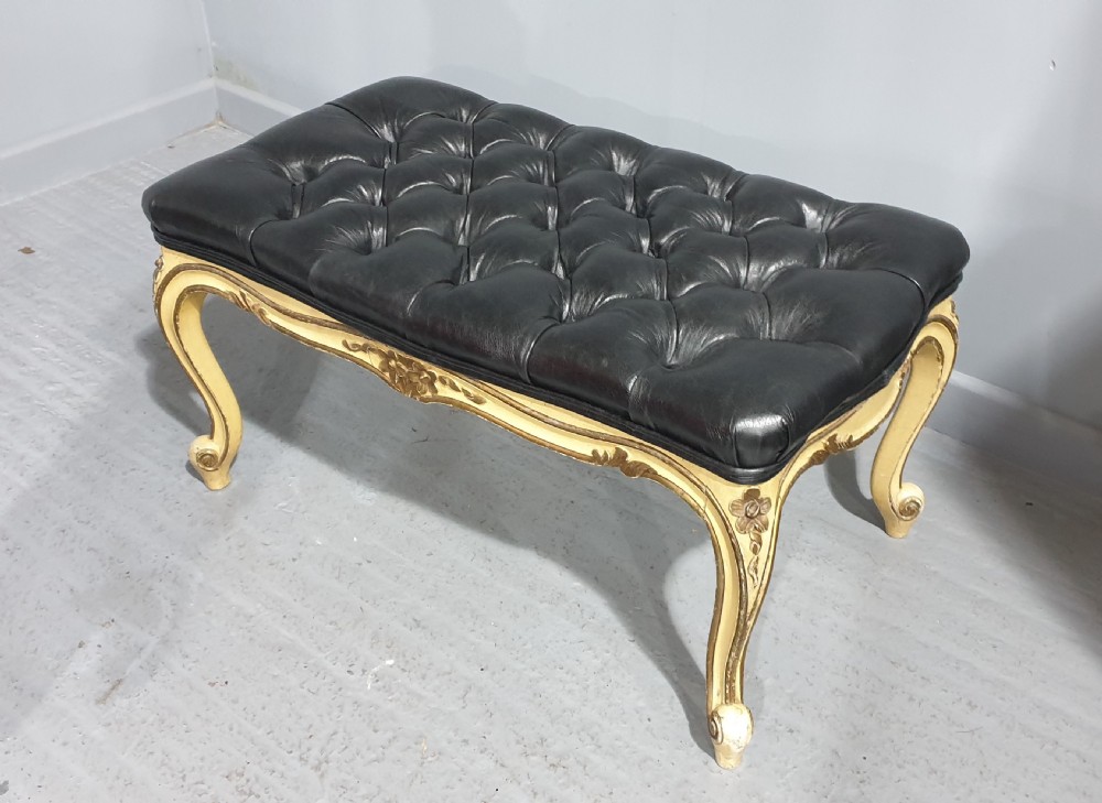 A long rectangular stool featuring a black buttoned leather tufted seat set upon a carved cream and gold painted wooden frame with cabriole legs. View VIII