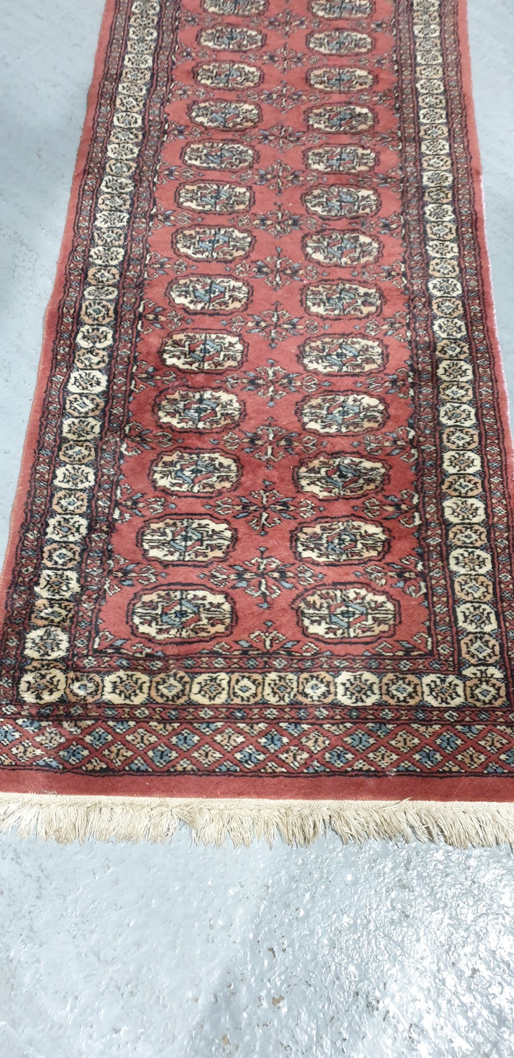 An extra-long hand-knotted wool carpet runner featuring repeated geometric medallion patterns on a terracotta red field with patterned cream borders. View III