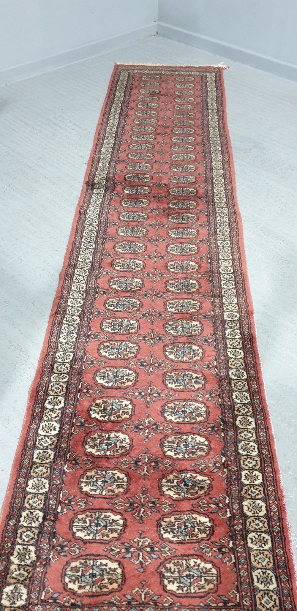 An extra-long hand-knotted wool carpet runner featuring repeated geometric medallion patterns on a terracotta red field with patterned cream borders. View V