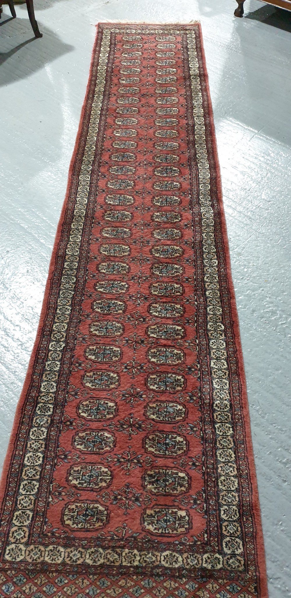 An extra-long hand-knotted wool carpet runner featuring repeated geometric medallion patterns on a terracotta red field with patterned cream borders. View VI