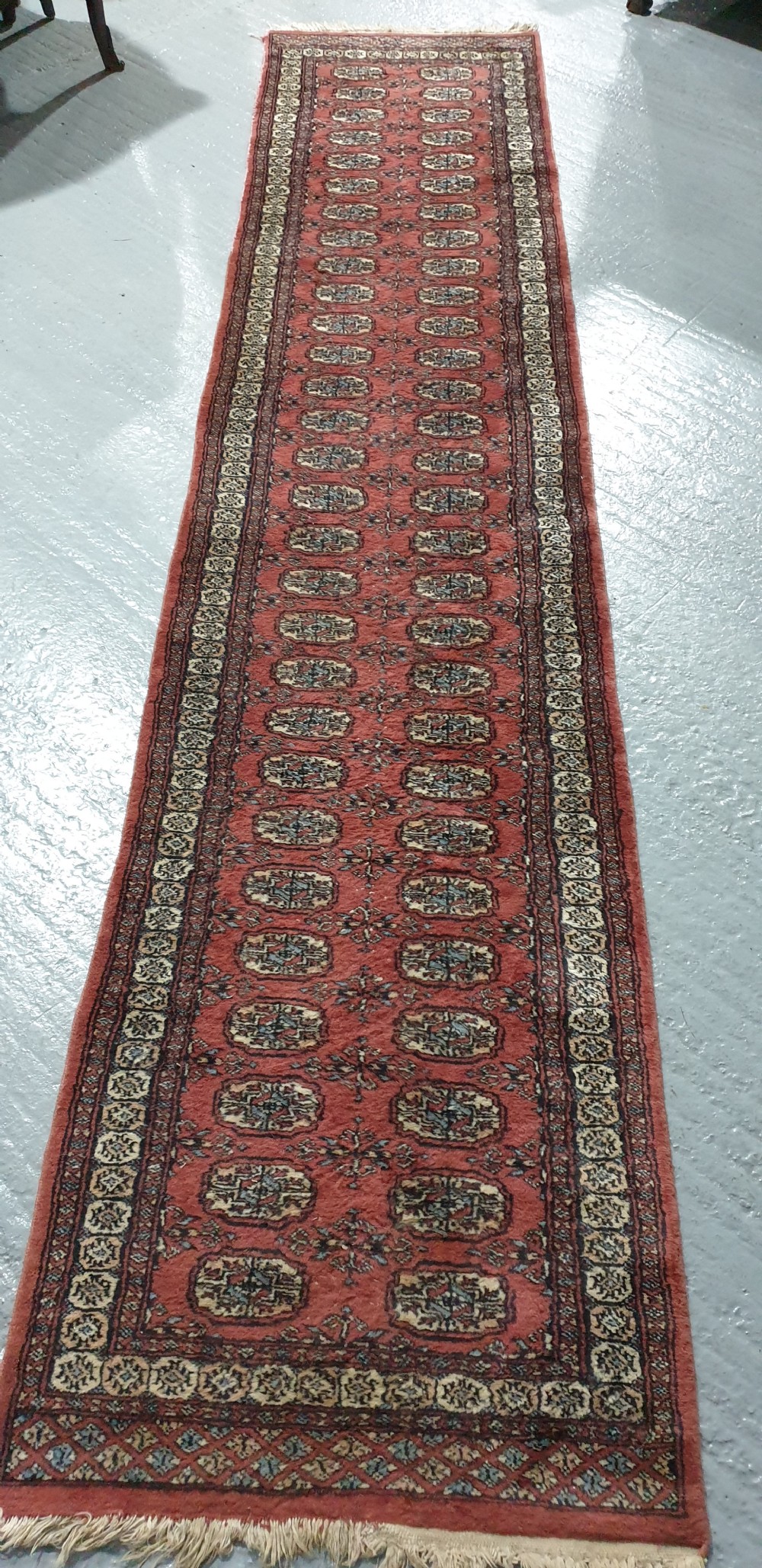 An extra-long hand-knotted wool carpet runner featuring repeated geometric medallion patterns on a terracotta red field with patterned cream borders. View VII