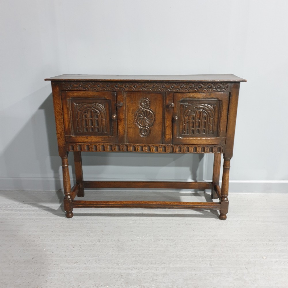 A small dark oak carved sideboard with two decorative panelled doors, turned legs, and lower stretchers.