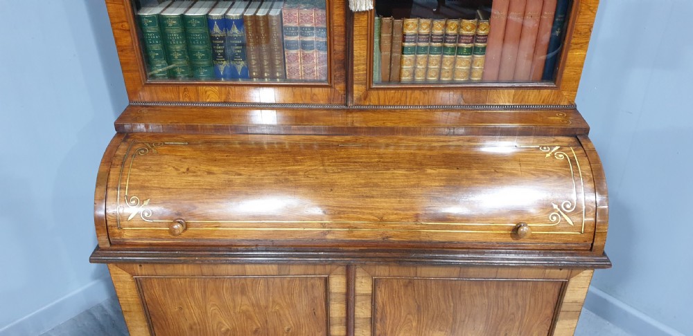 A Regency rosewood bureau bookcase featuring a scrolled pediment with brass finials, glazed upper display doors, a roll-top cylinder writing section with brass inlay, and a twin-door lower cupboard. View V