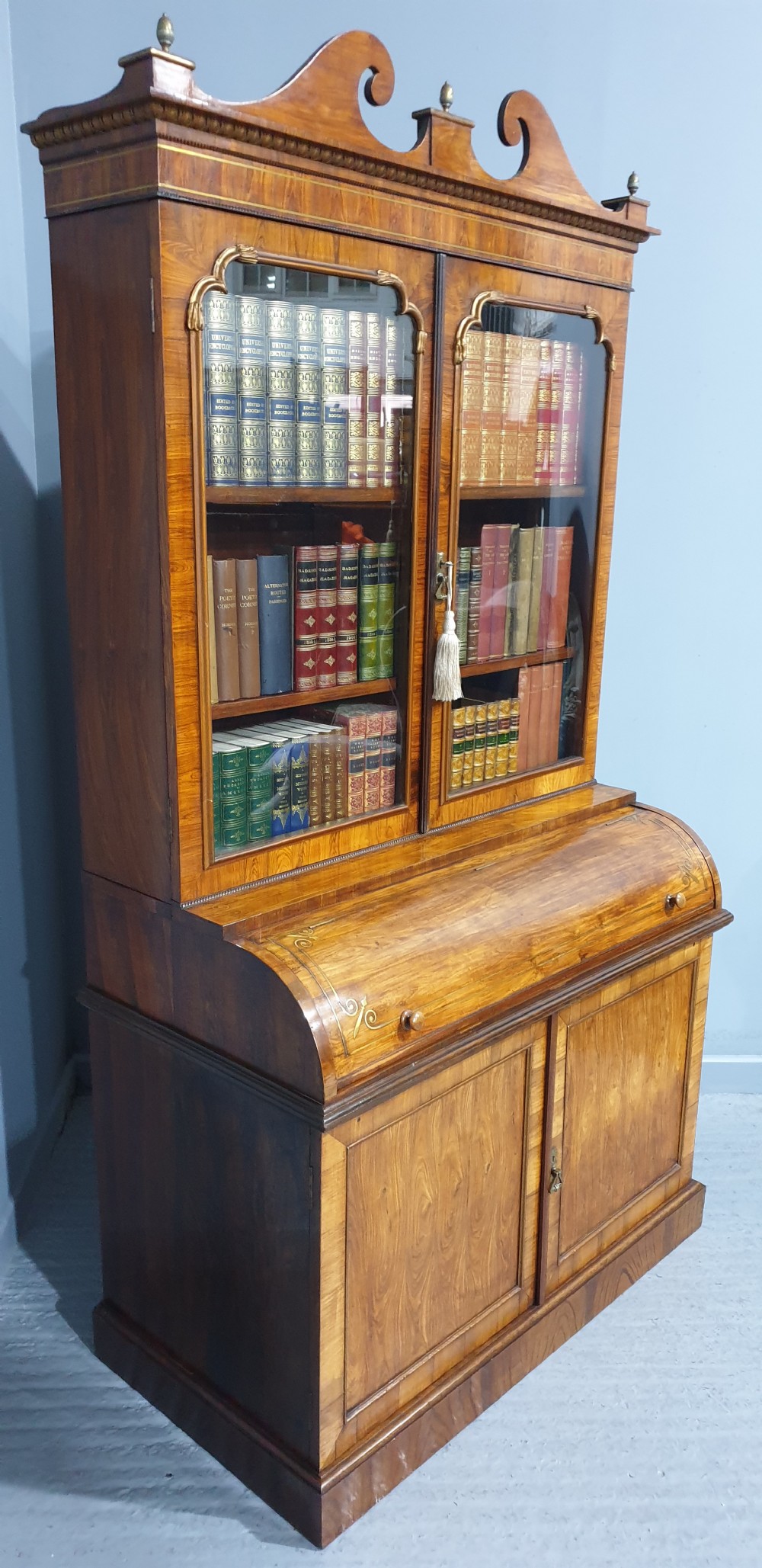 A Regency rosewood bureau bookcase featuring a scrolled pediment with brass finials, glazed upper display doors, a roll-top cylinder writing section with brass inlay, and a twin-door lower cupboard. View VII