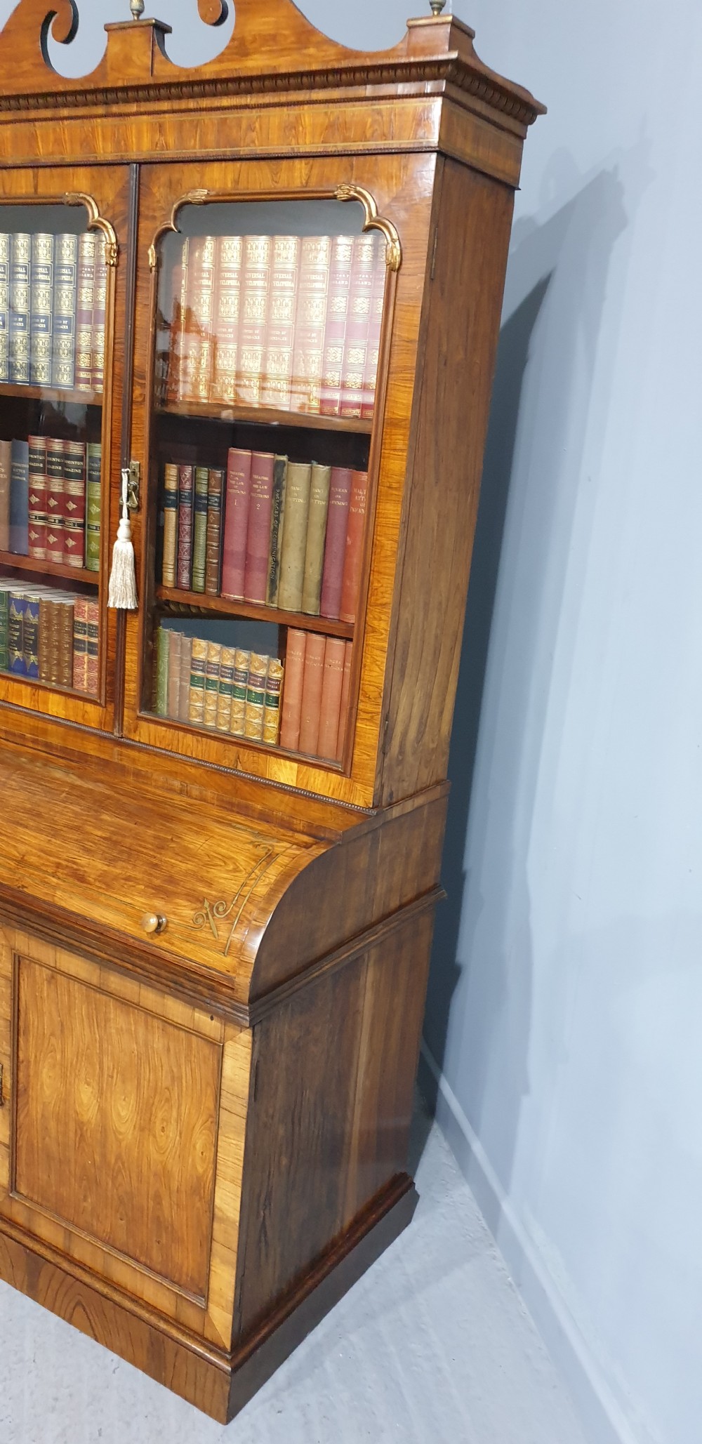 A Regency rosewood bureau bookcase featuring a scrolled pediment with brass finials, glazed upper display doors, a roll-top cylinder writing section with brass inlay, and a twin-door lower cupboard. View VIII
