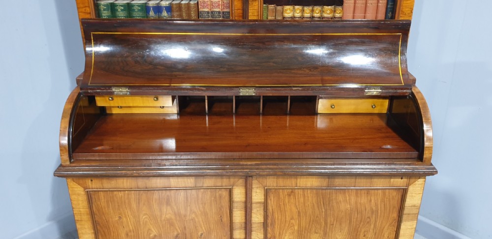 A Regency rosewood bureau bookcase featuring a scrolled pediment with brass finials, glazed upper display doors, a roll-top cylinder writing section with brass inlay, and a twin-door lower cupboard. View IX