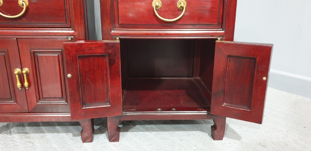 A matching pair of reddish-brown lacquered Oriental bedside cabinets featuring brass handles, single upper drawers, double cupboard doors, and panelled detailing. View VI
