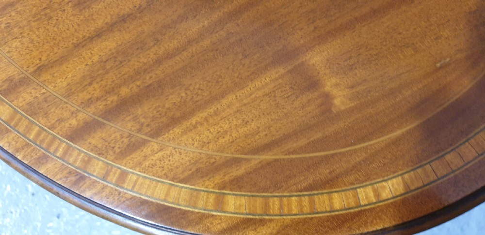 An Edwardian mahogany baby Sutherland drop-leaf table featuring an oval top with satinwood crossbanding, central marquetry patera, slender turned legs, and brass casters. View V
