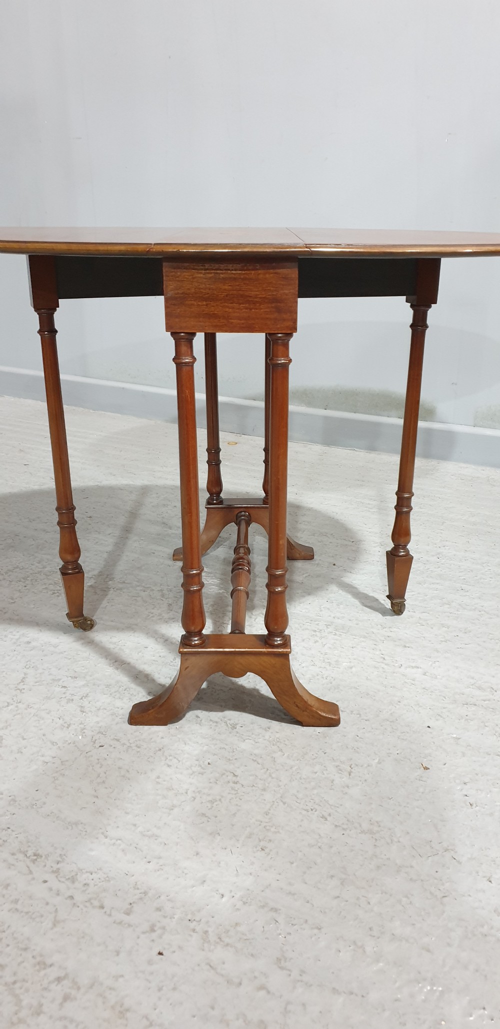 An Edwardian mahogany baby Sutherland drop-leaf table featuring an oval top with satinwood crossbanding, central marquetry patera, slender turned legs, and brass casters. View VIII