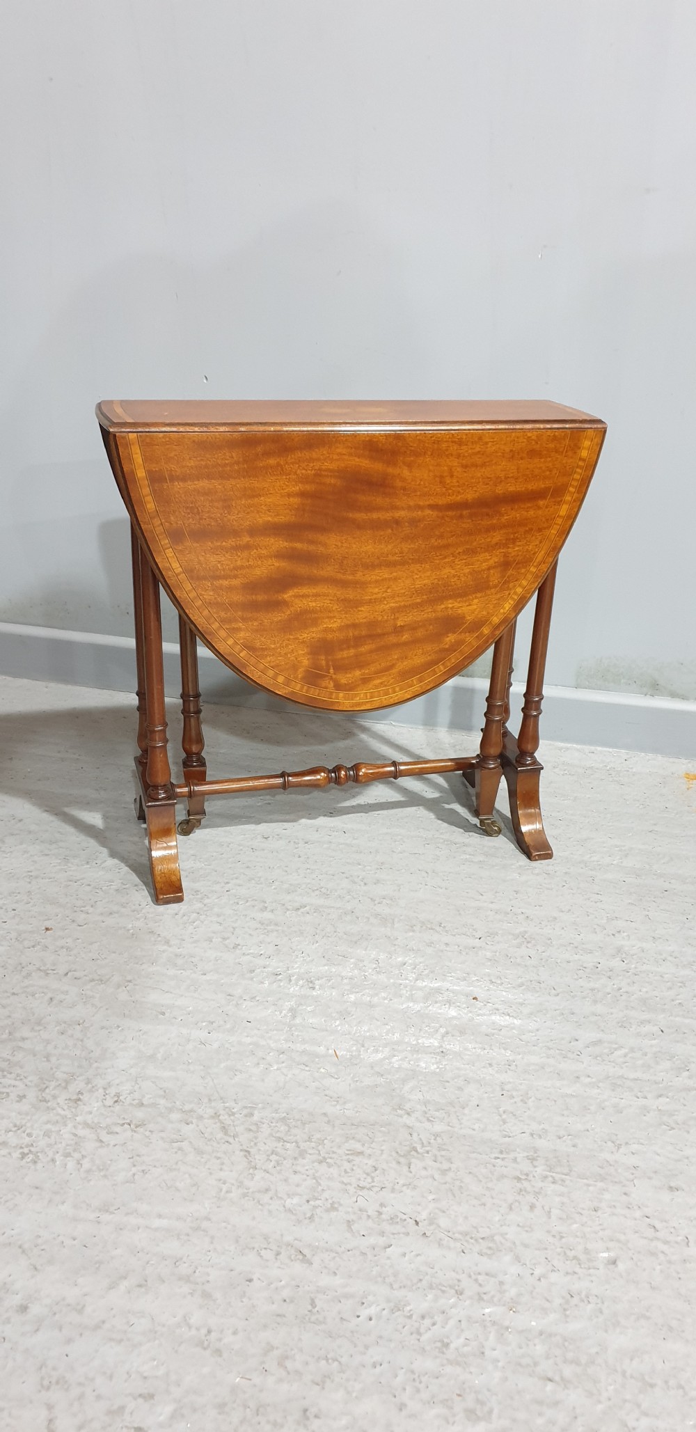 An Edwardian mahogany baby Sutherland drop-leaf table featuring an oval top with satinwood crossbanding, central marquetry patera, slender turned legs, and brass casters. View IX
