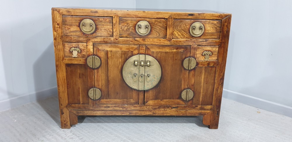 A 19th-century Chinese elm sideboard featuring three upper drawers, two panelled cupboard doors with a central circular brass lockplate, and additional small side drawers with brass fittings. View II