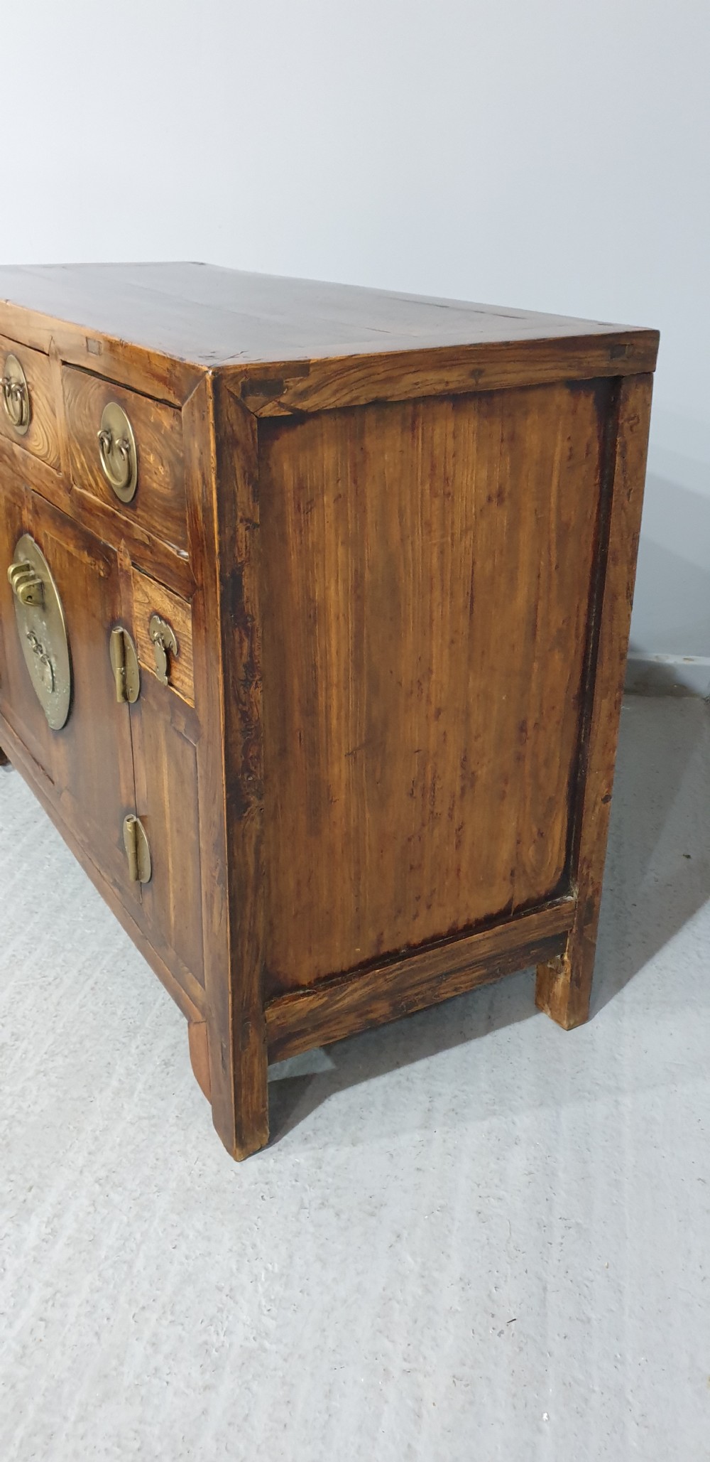 A 19th-century Chinese elm sideboard featuring three upper drawers, two panelled cupboard doors with a central circular brass lockplate, and additional small side drawers with brass fittings. View V