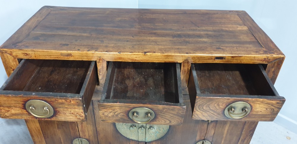 A 19th-century Chinese elm sideboard featuring three upper drawers, two panelled cupboard doors with a central circular brass lockplate, and additional small side drawers with brass fittings. View VII