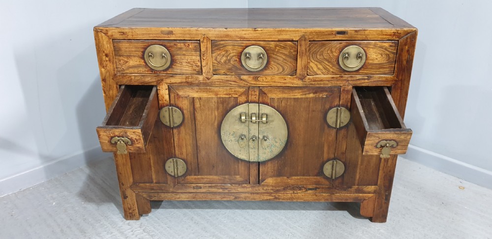 A 19th-century Chinese elm sideboard featuring three upper drawers, two panelled cupboard doors with a central circular brass lockplate, and additional small side drawers with brass fittings. View VIII