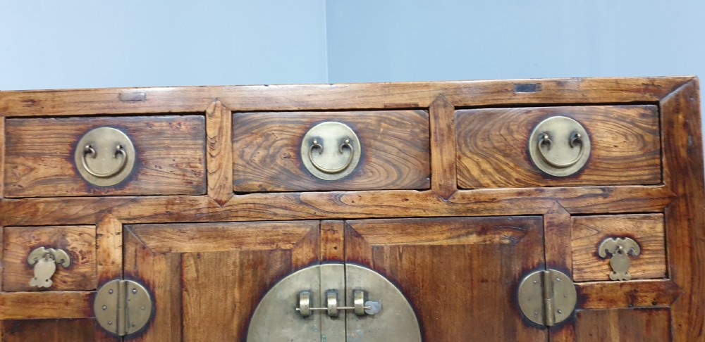A 19th-century Chinese elm sideboard featuring three upper drawers, two panelled cupboard doors with a central circular brass lockplate, and additional small side drawers with brass fittings. View XII
