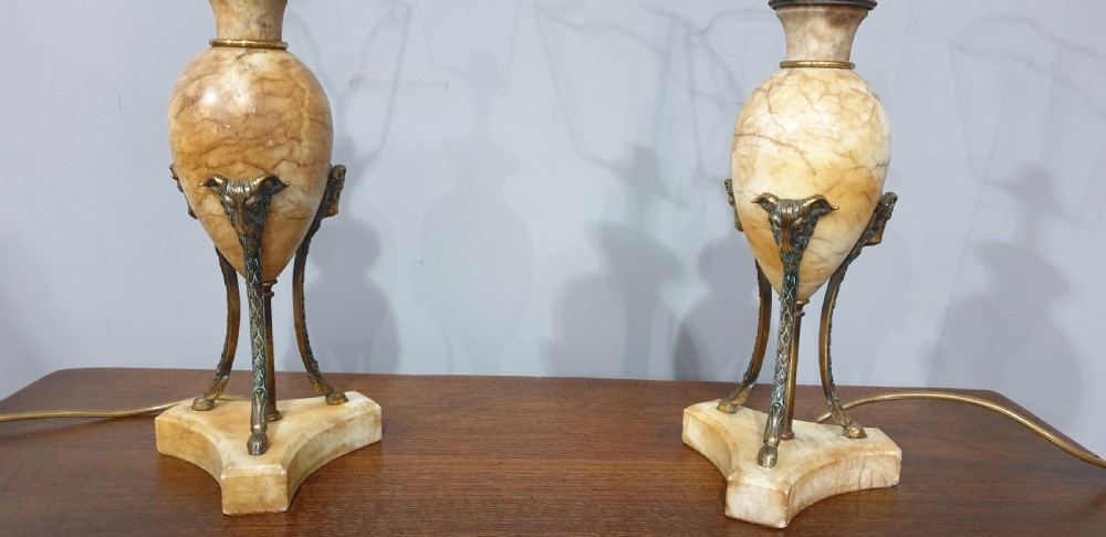 A pair of urn-form alabaster table lamps with brass tripod supports and pleated cream fabric shades resting on a wooden surface. View IV