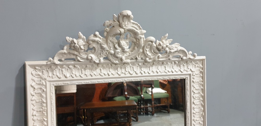 A rectangular off-white painted wall mirror featuring a top carved crest with leaf motifs and a decorative moulded frame. View IV