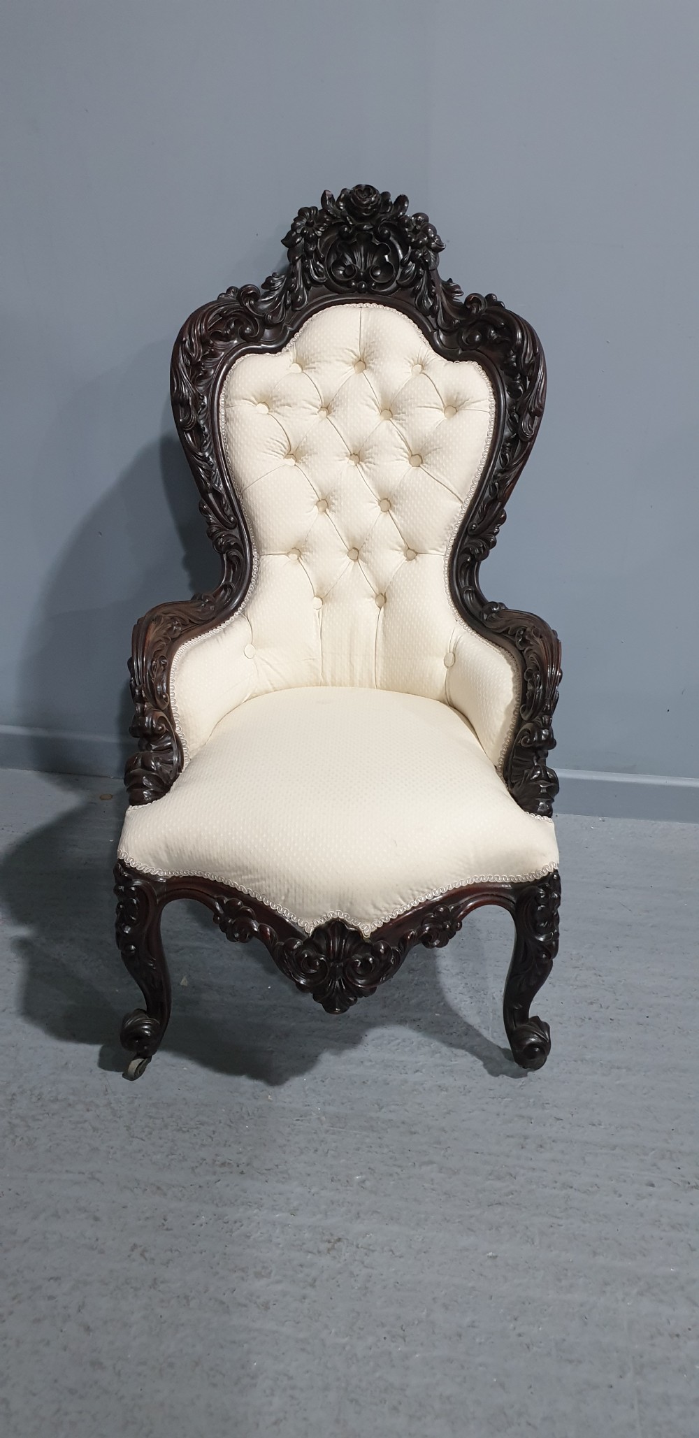 An antique Victorian dark mahogany armchair featuring an ornate floral carved crest, button-tufted cream upholstery, and carved cabriole legs on castors. View III