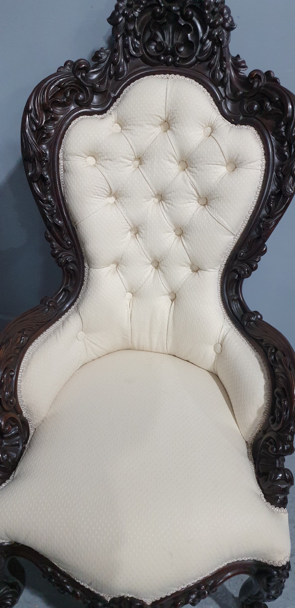 An antique Victorian dark mahogany armchair featuring an ornate floral carved crest, button-tufted cream upholstery, and carved cabriole legs on castors. View IV