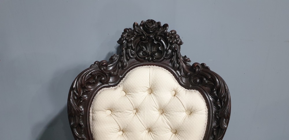 An antique Victorian dark mahogany armchair featuring an ornate floral carved crest, button-tufted cream upholstery, and carved cabriole legs on castors. View VI