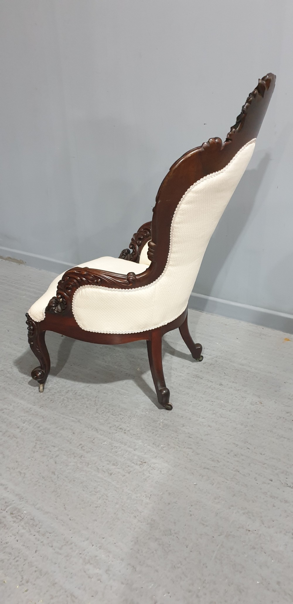 An antique Victorian dark mahogany armchair featuring an ornate floral carved crest, button-tufted cream upholstery, and carved cabriole legs on castors. View VII