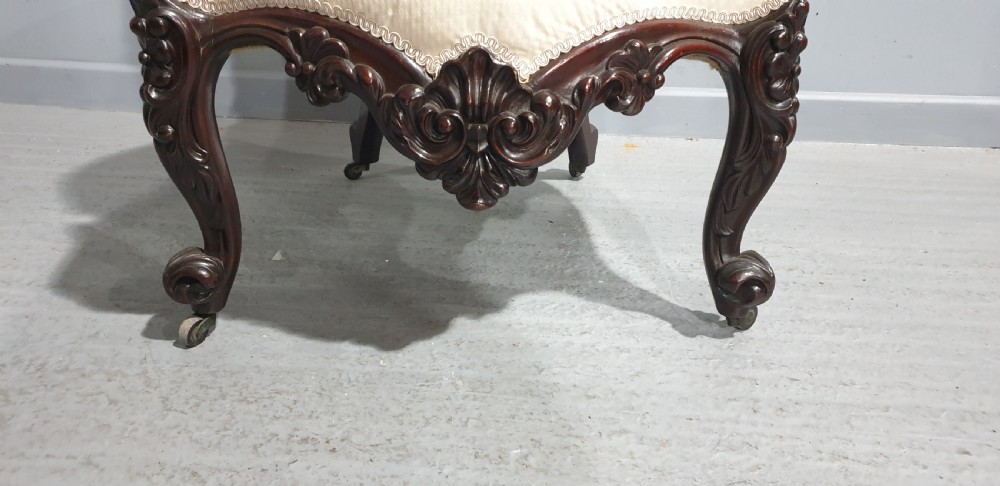 An antique Victorian dark mahogany armchair featuring an ornate floral carved crest, button-tufted cream upholstery, and carved cabriole legs on castors. View VIII