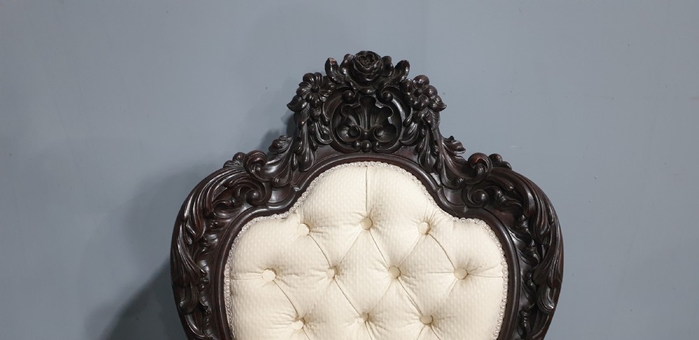An antique Victorian dark mahogany armchair featuring an ornate floral carved crest, button-tufted cream upholstery, and carved cabriole legs on castors. View IX