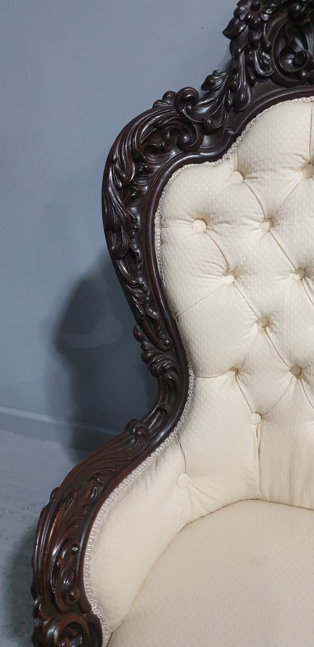 An antique Victorian dark mahogany armchair featuring an ornate floral carved crest, button-tufted cream upholstery, and carved cabriole legs on castors. View X