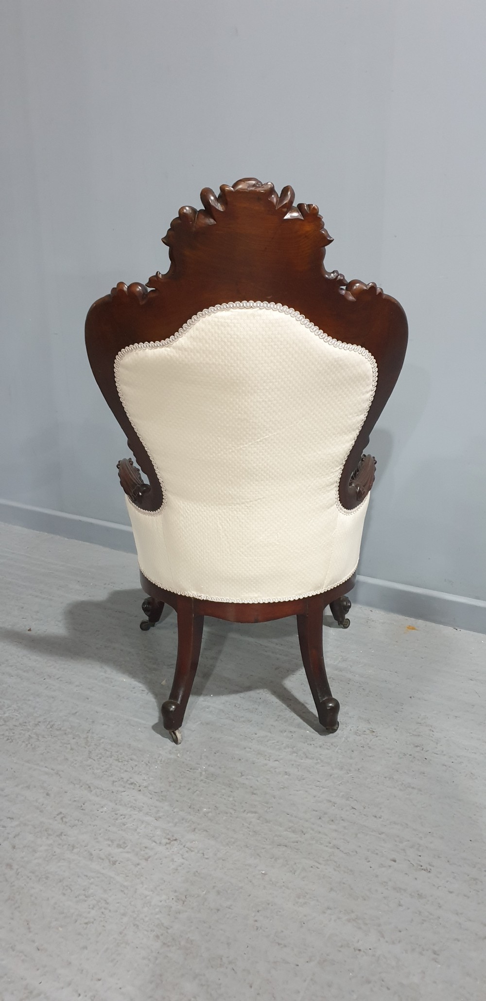 An antique Victorian dark mahogany armchair featuring an ornate floral carved crest, button-tufted cream upholstery, and carved cabriole legs on castors. View XI