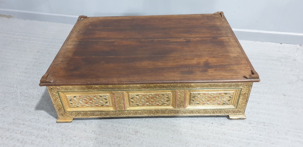 A rectangular low wooden stool featuring a plain dark timber top surface above carved, gilded side panels with colourful geometric inlays. View II