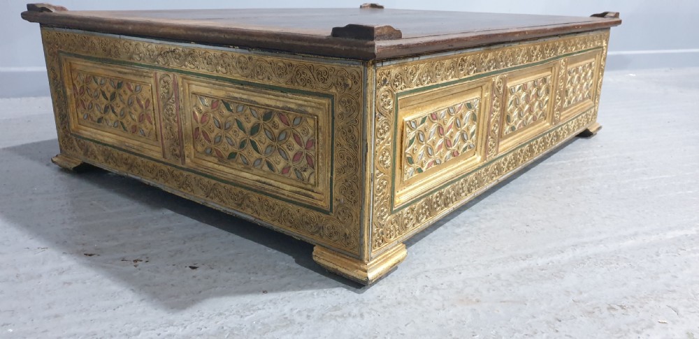 A rectangular low wooden stool featuring a plain dark timber top surface above carved, gilded side panels with colourful geometric inlays. View III