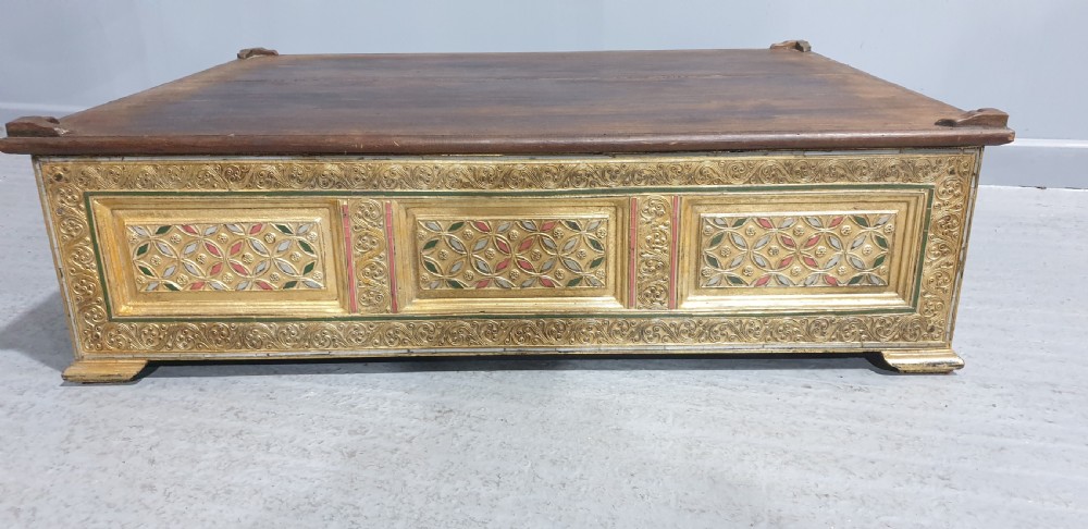 A rectangular low wooden stool featuring a plain dark timber top surface above carved, gilded side panels with colourful geometric inlays. View V