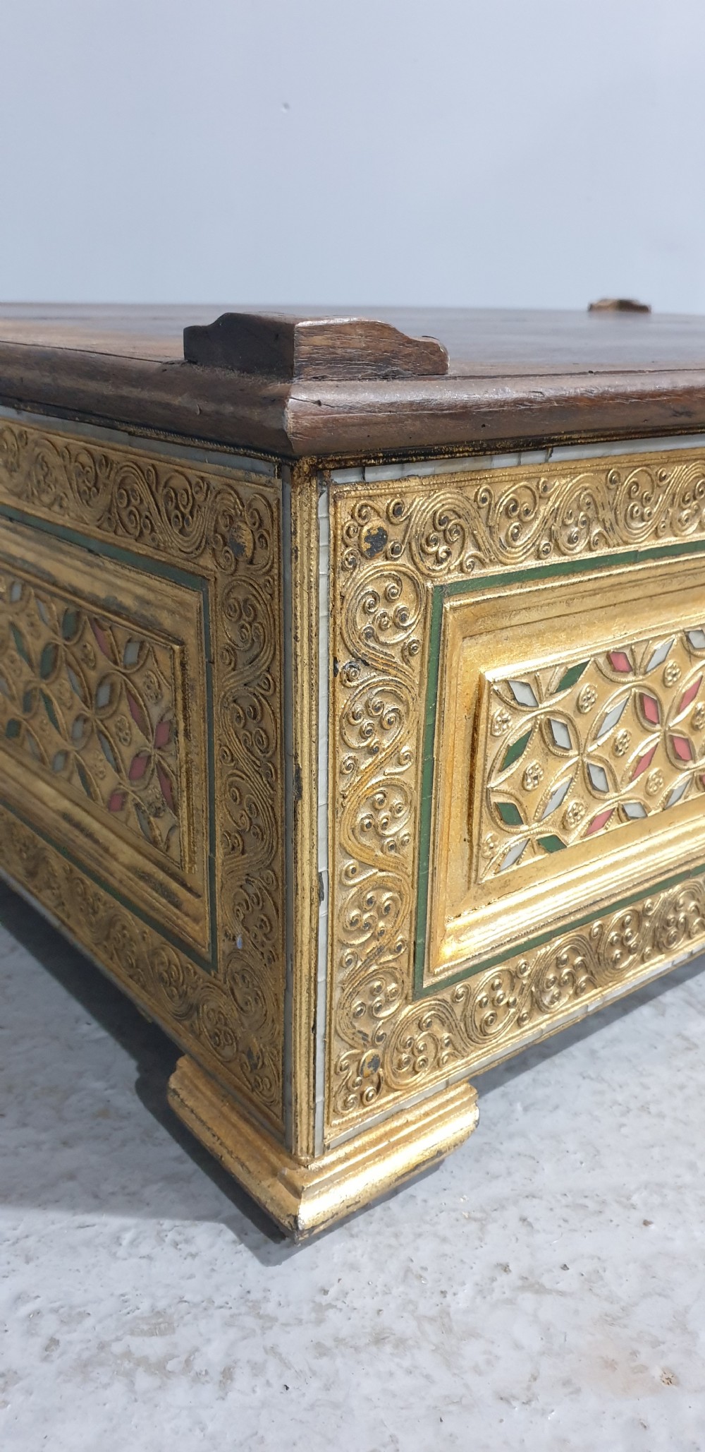 A rectangular low wooden stool featuring a plain dark timber top surface above carved, gilded side panels with colourful geometric inlays. View VI