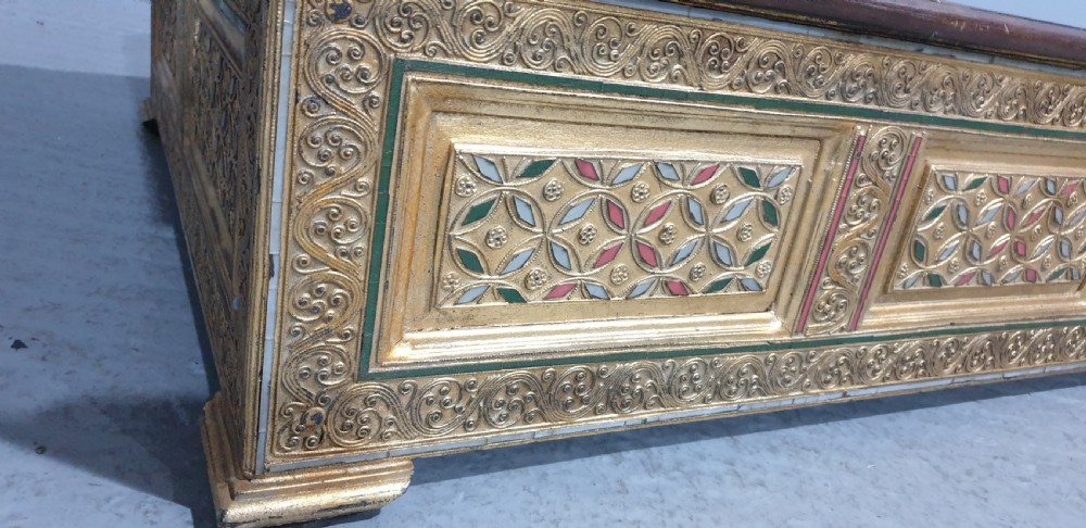 A rectangular low wooden stool featuring a plain dark timber top surface above carved, gilded side panels with colourful geometric inlays. View VII