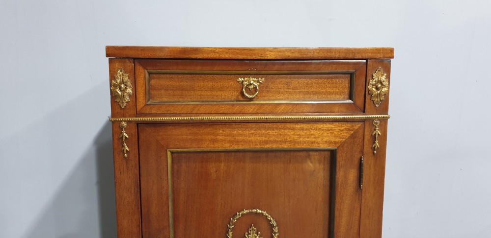 An antique French Empire style mahogany side cabinet featuring a single top drawer, a lower cupboard door decorated with a brass laurel wreath mount, and square tapered legs with brass feet. View II