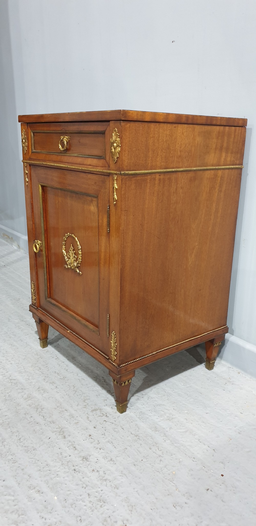 An antique French Empire style mahogany side cabinet featuring a single top drawer, a lower cupboard door decorated with a brass laurel wreath mount, and square tapered legs with brass feet. View VI