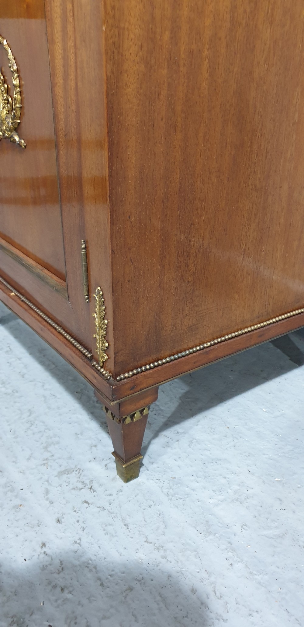An antique French Empire style mahogany side cabinet featuring a single top drawer, a lower cupboard door decorated with a brass laurel wreath mount, and square tapered legs with brass feet. View VII