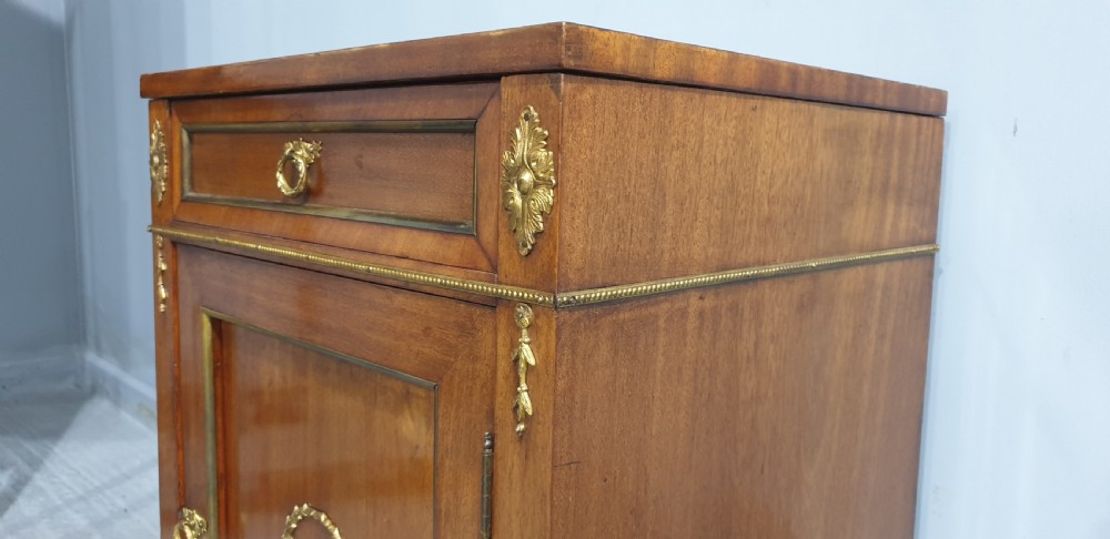 An antique French Empire style mahogany side cabinet featuring a single top drawer, a lower cupboard door decorated with a brass laurel wreath mount, and square tapered legs with brass feet. View IX