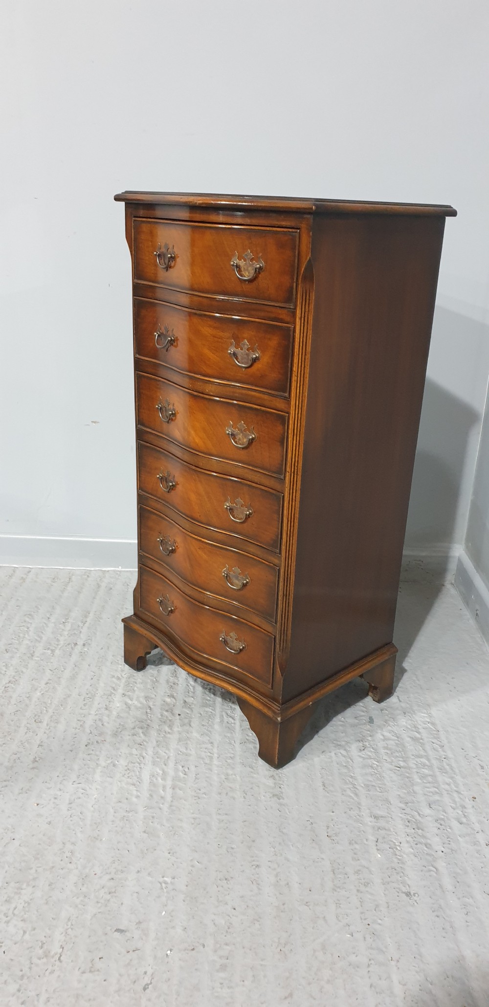 A tall, narrow mahogany chest of drawers featuring a serpentine front with six drawers and decorative brass drop handles. View II
