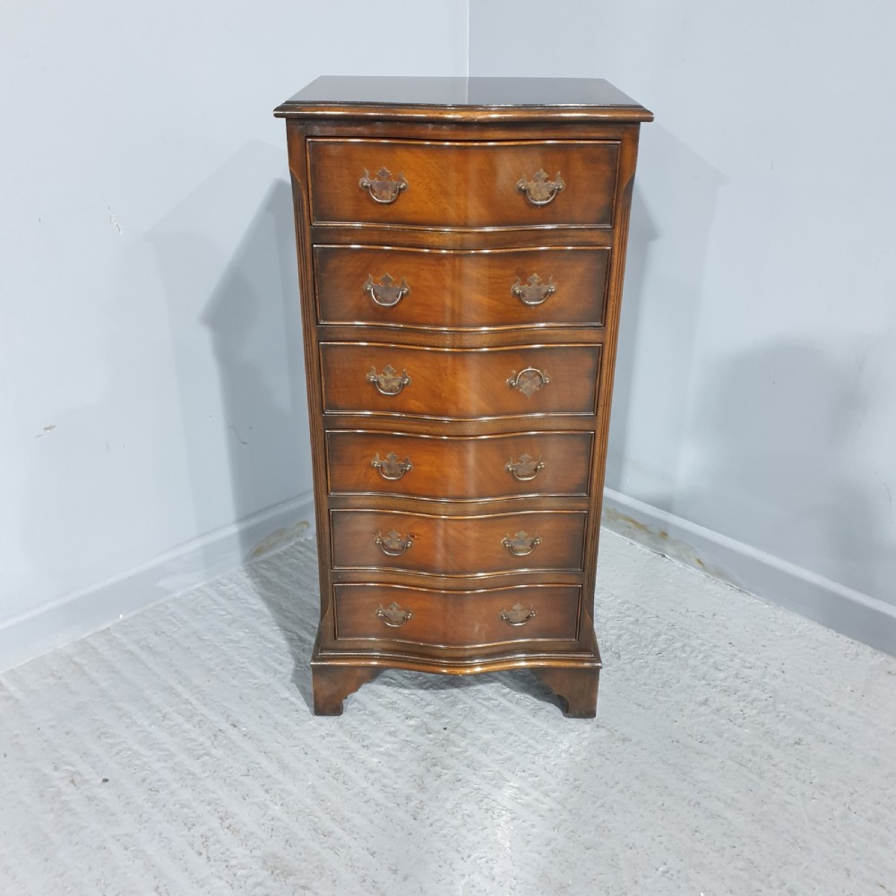 A tall, narrow mahogany chest of drawers featuring a serpentine front with six drawers and decorative brass drop handles. View IV