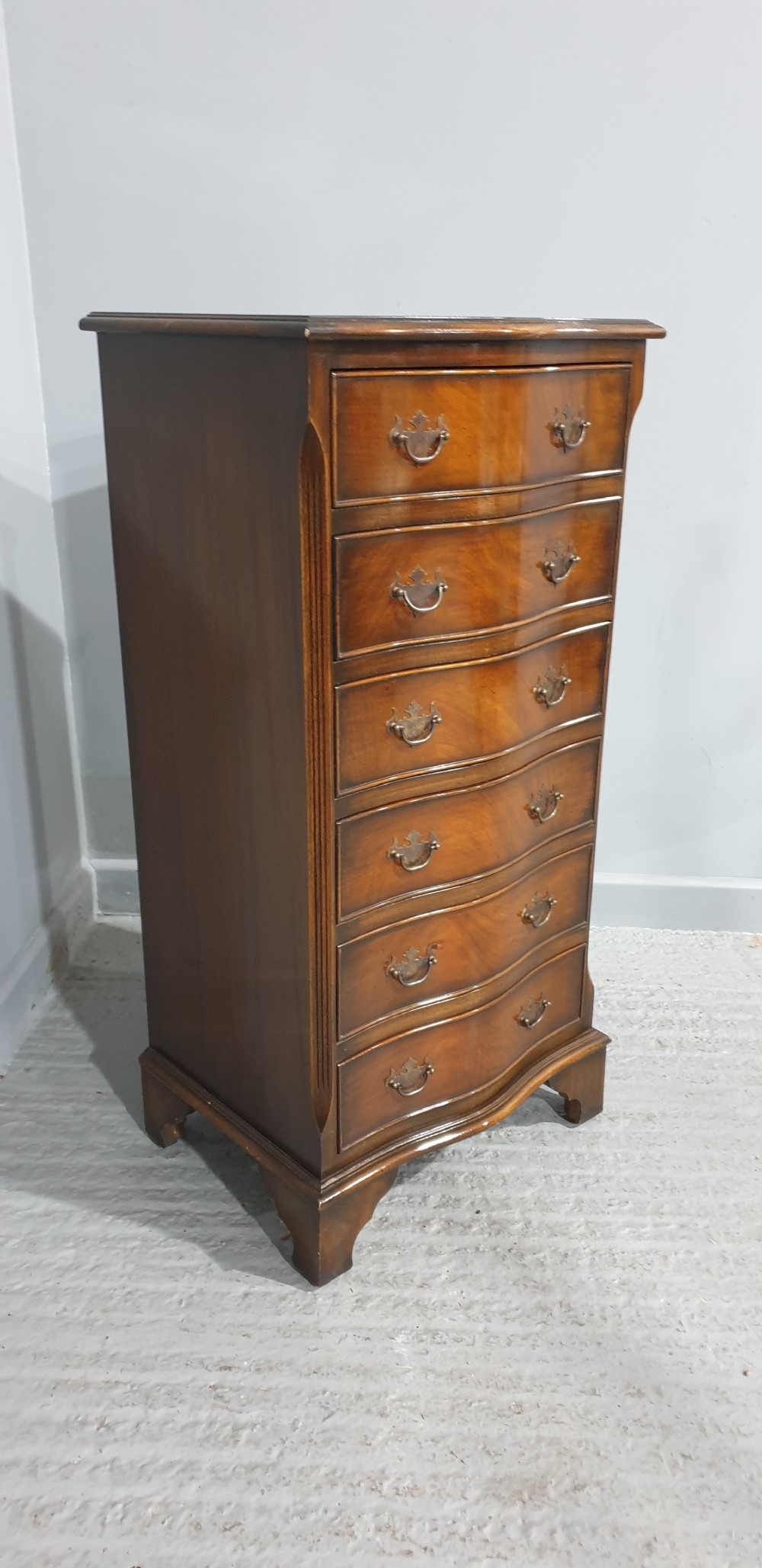 A tall, narrow mahogany chest of drawers featuring a serpentine front with six drawers and decorative brass drop handles. View VI