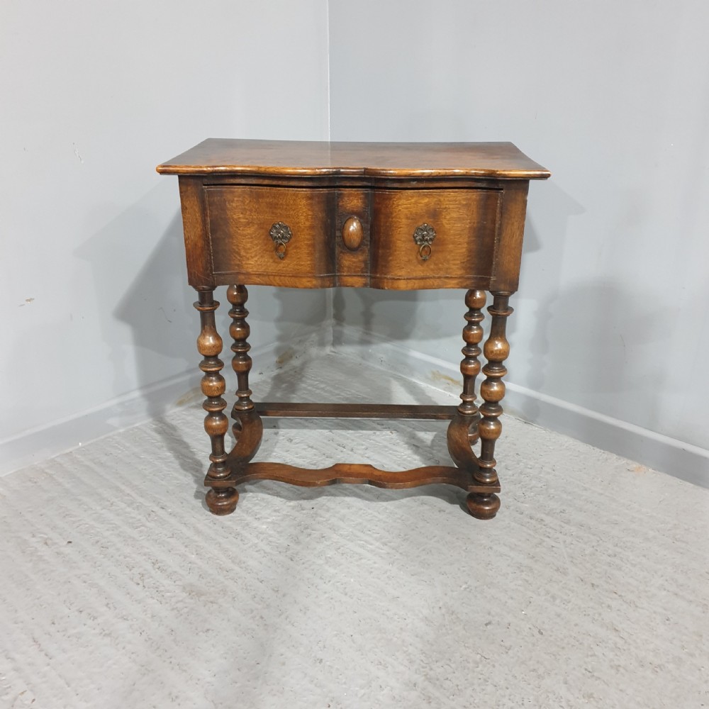 An antique oak side table featuring bobbin turned legs, a shaped frieze drawer with brass drop handles, and a wavy cross-stretcher base.