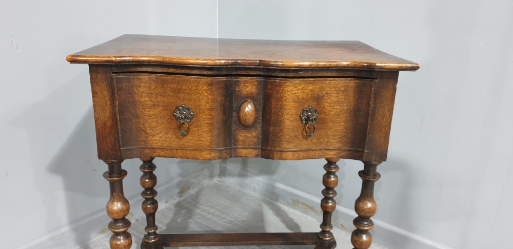 An antique oak side table featuring bobbin turned legs, a shaped frieze drawer with brass drop handles, and a wavy cross-stretcher base. View II