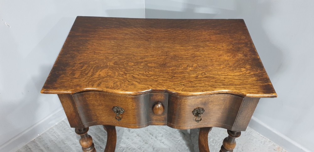 An antique oak side table featuring bobbin turned legs, a shaped frieze drawer with brass drop handles, and a wavy cross-stretcher base. View IV