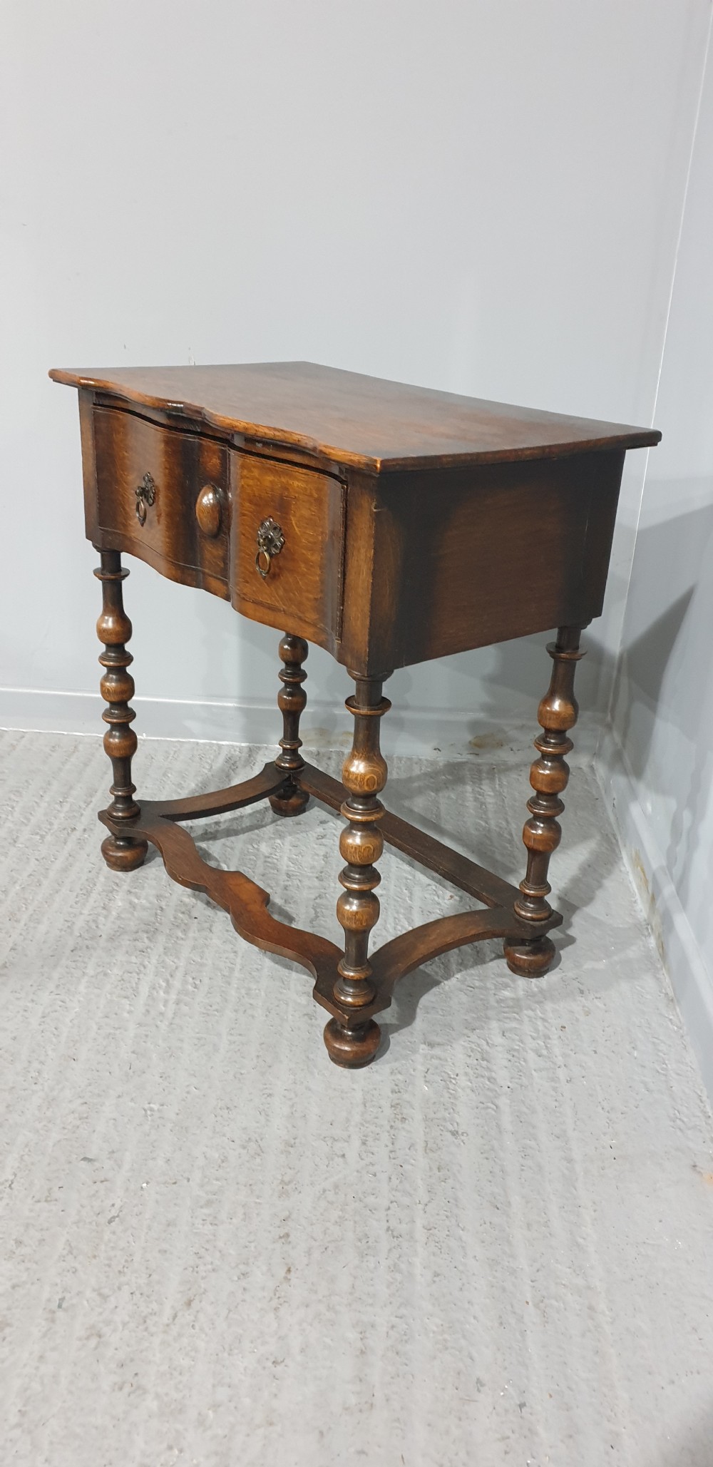 An antique oak side table featuring bobbin turned legs, a shaped frieze drawer with brass drop handles, and a wavy cross-stretcher base. View V